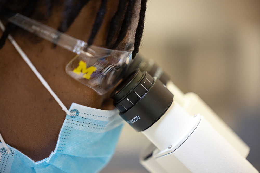 A closeup of a University of Michigan student wearing a mask looking through a microscope