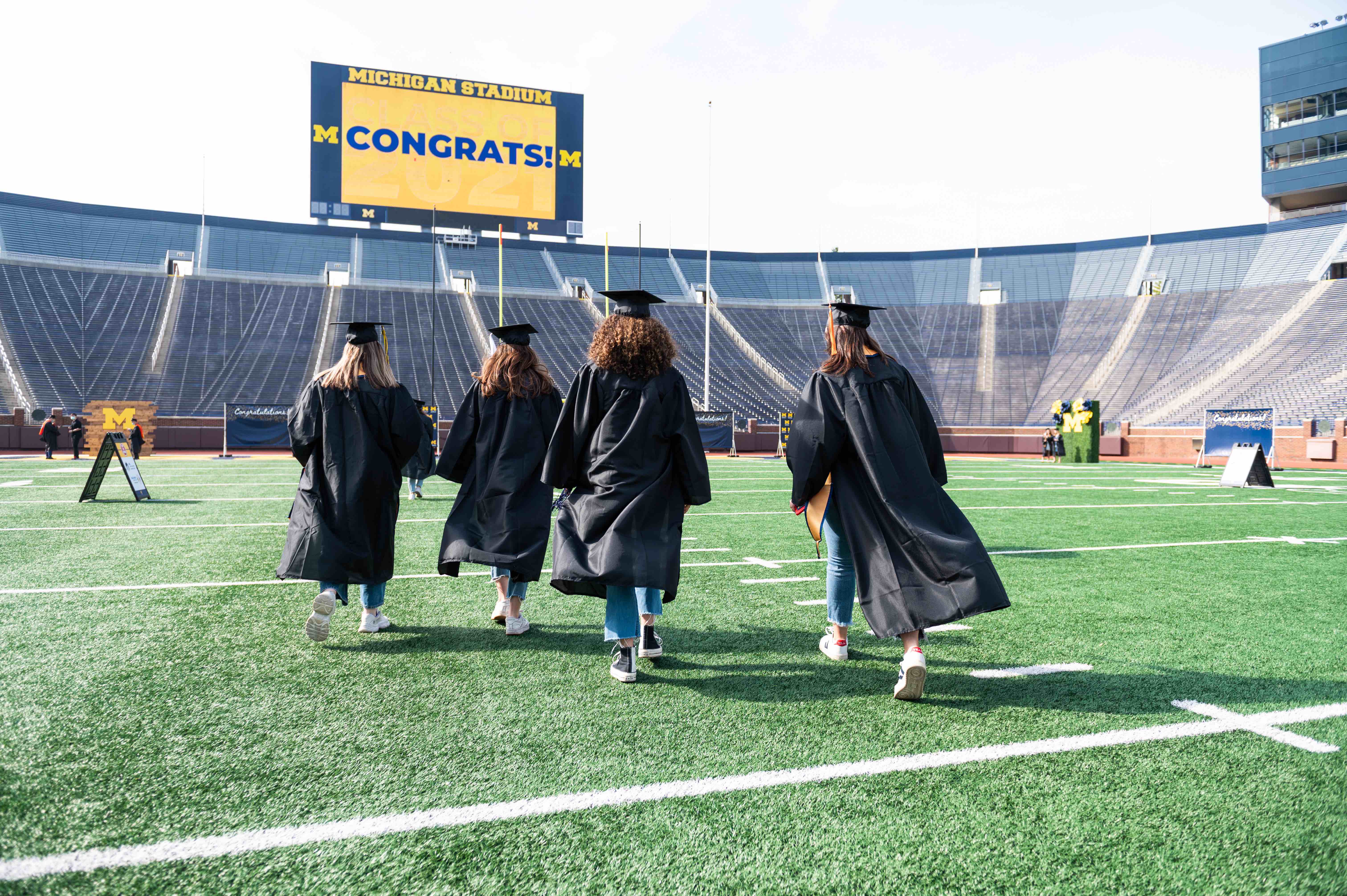 Four University of Michigan graduates wearing caps and gowns walk across the field at Michigan Stadium